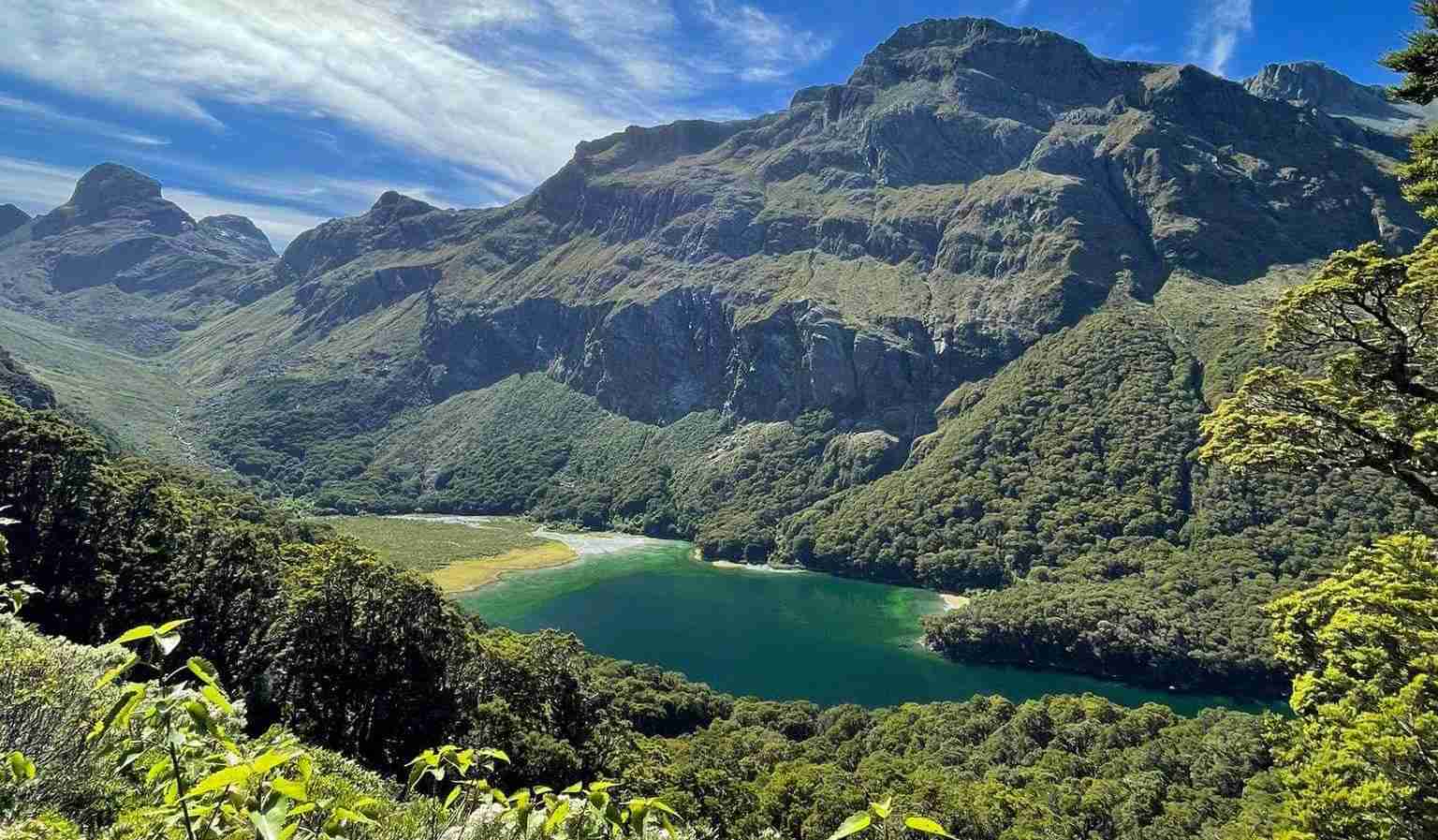 A breathtaking view of the Routeburn Track, showcasing the pristine lake surrounded by lush greenery and towering mountains under a clear blue sky.